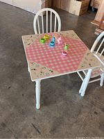 Kids wooden tea table with pink gingham pattern top and floral border, with two chairs and colorful plastic toy tea set placed on table