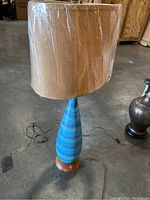 Blue ceramic table lamp with large brown fabric shade wrapped in plastic, electrical cord visible on floor