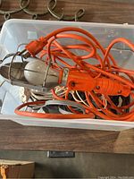 Mechanic light with metal cage, orange handle, and attached orange power cord coiled inside a plastic bin.