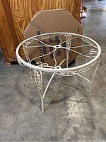 White round iron patio table with decorative scrollwork on top and legs, placed on concrete floor.