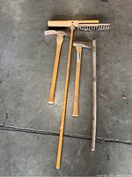 Photo showing four yard tools laid on the floor: garden rake, splitting maul, pick axe, and push broom. All tools show scratches and signs of use.
