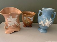 Front view of three Weller Pottery vases: rose/pink Cameo, brown footed vase with blossoms and butterfly, and blue Cameo pattern.
