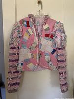 Front view of a bolero style jacket hanging on a wall. The jacket is handmade with multi-colored patchwork quilted body and long dolman style sleeves made of crocheted fabric strips. It has ruffled trim around the neckline and front edges, with button closures.