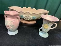 Three vintage Hull pottery pieces displayed on black cloth with green curtain background: large bowl, cornucopia vase, and floral vase with handle.
