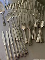 Photo showing various silverplate flatware pieces including knives, forks, spoons, and serving utensils arranged on a surface. The majority are Oneida Churchill pattern with beaded edges, alongside assorted pieces from other patterns.