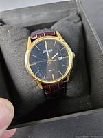 Photo showing the Citizen men's quartz watch with black dial, gold-tone case, and brown leather strap placed on black cushion inside box.