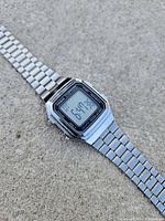 Front angle of the Casio digital watch showing the LCD display indicating time, day and date. Stainless steel casing and band visible.