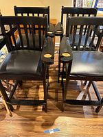 Four black wooden bar stools with padded black seats and cup holders on armrests arranged together on wooden floor.