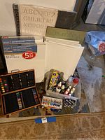 Overall group shot showing canvases, stencil sheets, wooden colored pencil case and plastic bin with assorted paint bottles and tubes.