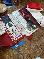 Photo showing multiple Christmas-themed table runners with snowmen, Christmas trees, plaid, and sleigh patterns, along with red placemats and folded linens on a carpet.