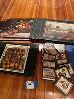 Image showing cork placemats with various printed designs including basket of apples, ducks, and a tiered fruit design, alongside two sets of coasters with fruit and ship illustrations.