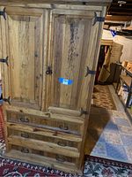 Front view of a light wood TV cabinet with distressed finish, two large doors with iron hinges, and three drawers at the bottom with iron handles. Cabinet stands on a rug in a basement.