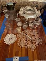 Photo showing full set including punch bowl, six mugs, crystal decanter with stopper, large crystal pitcher with inner ice holder (cracked), silver punch ladle, and two lace doilies placed on wood floor.
