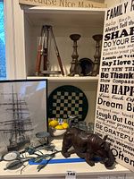 Framed ship art featuring a tall sailing ship, wood signs with text, metal candlestick holders, glass chess board, metal hippo statue, metal bowl with faux lemons, and green metal bird candle holder.