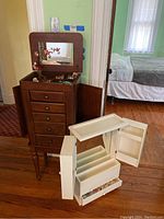 Full view of the tall brown wooden jewelry chest standing on four legs beside the white jewelry wall cabinet. The top lid of the chest is open showing compartments and the mirror. The white wall cabinet is detached and placed beside the chest with side doors open exposing inner compartments and drawers visible at front.