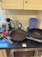 Wide shot of all items on countertop showing French press, carafe, frying pan, utensils, plastic pitchers, stock pot.