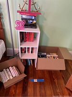 Photo shows white 2-shelf bookcase filled with books, two cardboard boxes of more books beside it, and a pink butterfly bookend on the top shelf with gold metal anchor bookends on stacked books above.