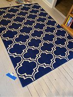 Full view of rectangular navy blue rug with white repeating geometric quatrefoil motif on light wood floor near bookshelf.