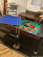 Gaming table with pool table surface showing balls, pool cues in metal holder, orange ping pong paddles on table, and blue table tennis top partially placed on table.