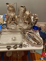 Full view of lot including glass serving dish, silver plated oval tray, multiple silver plated pitchers, coffee pot, teapot, crystal dishes, and five napkin rings laid in front.