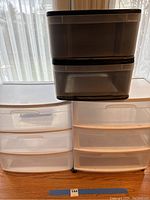 Two white three-drawer plastic storage bins with castors and one black two-drawer plastic storage bin stacked on floor.