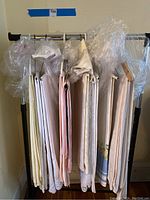 Hanging view of 11 tablecloths on rack, wrapped plastic wrap mostly around hangers, showing various lengths and colors mostly off-white and cream.
