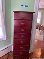 Front view of tall dresser showing seven drawers with brass handles against a light green wall.