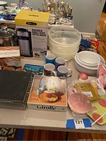 Photo showing boxed Bella Coffee Maker, plastic bowls, mugs, cake mold, birthday cupcake dish and other assorted kitchen items on table