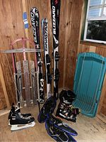Wooden sled, plastic kids sled, 3 pairs of skis including Blizzard skis, snowshoes, Tecnica ski boots, and blue bindings visible in a wooden shed corner.