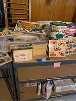 Wide view of various puzzles and toy containers including US map puzzle, Lincoln Logs tin and Tinkertoys tub on shelving