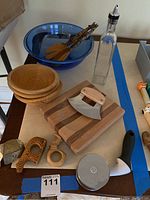 View of entire lot showing wooden bowls, blue glass bowl, salad lifters with zebra handles, oil/vinegar bottle, napkin rings, herb board and mezzaluna chopper, pizza cutter