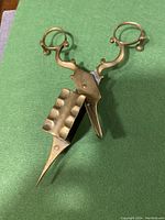 Photo of the brass candle snuffer scissors laid on green surface showing detail of scissor handles, wick trimmer box, and feet.