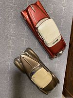 Top and angled view of both collectible toy cars on grey textured surface showing the red Cadillac convertible and metallic beige Buick