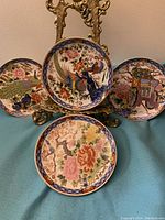 Four vintage hand-painted plates displayed on a decorative metal stand showing different traditional Japanese designs including birds, flowers, and peacocks.