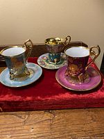 Front view of three antique demitasse cups each with their matching saucers, on a red velvet cloth. Cups show detailed gold floral decoration and unique colors (blue, magenta, and gold with scene).