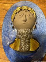 Close-up of the ceramic plaque showing the detailed 3D sculpted face with gold accents and textured collar on blue background.