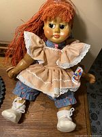 Front-facing photo of the Naber Kids collectible doll with red yarn hair sitting on a wooden surface, wearing tan dress and blue floral pants, white shoes visible.