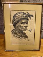 Framed artwork showing Indigenous man in profile wearing a woven hat with feather and smoking a pipe