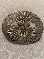 Front view of large oval silver brooch with floral design and central brown stone.