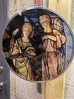 Front view of oval stained glass sun catcher showing two angels with musical instruments, framed in metal, hanging on a chain