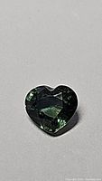 Close-up of a heart shaped blue-green sapphire with good clarity under lighting, viewed against a plain gray background.
