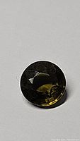 Close-up image of a round cut green tourmaline gemstone with dark olive-green hue and well-defined facets on a white background.