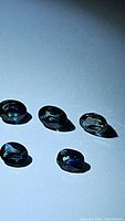Five oval faceted bi-colored sapphires in teal to dark blue hues on a light background, illuminated to show color and clarity.