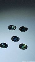 Five oval cut blue green sapphire gemstones arranged on a white background showing dimensions around 4x3mm each with blue and green hues.