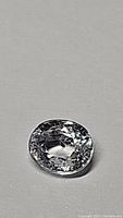 Close-up photo of a single oval faceted clear to very light blue sapphire gemstone on a neutral background.