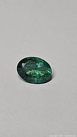 Close-up images of a single oval cut deep green Zambian emerald gemstone on a plain background, showing natural inclusions and facet cuts.