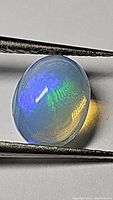 Oval cabochon cut natural untreated Ethiopian opal gemstone held by tweezers, showing blue and green flashes under light