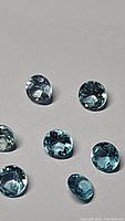 Close-up of seven loose round Paraiba blue Apatite gemstones on a grey surface, showing their diamond cut facets and vivid blue color.