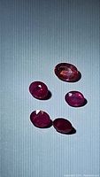 Five oval shape faceted pink and red gemstones displayed on light background under directional light, showing color and clarity.