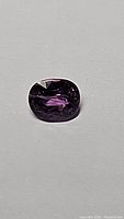 Purple sapphire gemstone from Madagascar shown on a grey surface, highlighting the deep purple color and oval cushion shape, with visible internal inclusions.